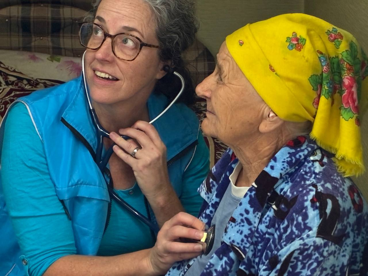 A GCF medical volunteer uses a stethoscope while caring for an elderly woman wearing a yellow floral headscarf.