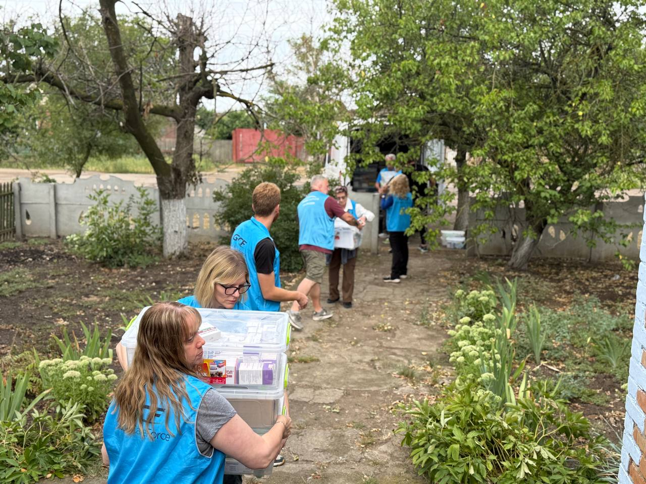 GCF volunteers carry clear medical supply bins together along a village path.